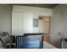 Schreibtisch in einem modernen Büro mit Computerbildschirm im Vordergrund. Im Hintergrund befinden sich weiße Schiebetüren, eine offene Zimmertür und ein grauer Sichtbeton-Deckenbereich.