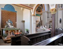 Innenansicht einer Kirche mit dekorativer Architektur. Im Vordergrund steht eine Pietà-Statue vor einer reich verzierten Rückwand mit brennenden Kerzen, während im Hintergrund der Altarbereich mit Säulen und einer Kanzel zu sehen ist.