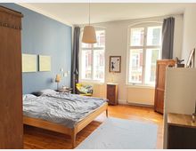 Schlafzimmer mit Holzboden, blauer Wand, einem Holzbett mit grauer Bettwäsche und einem gelben Sessel. Große helle Fenster lassen Tageslicht herein, umgeben von Holzmöbeln wie Kleiderschränken und Beistelltischen.