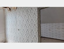Ein leerer, vernachlässigter Innenraum mit gemusterter Tapete an den Wänden, die teils beschädigt ist. Der Boden ist staubig, und im Hintergrund ist ein weiteres Zimmer mit ähnlicher abgenutzter Tapete sichtbar.