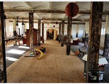 Großer, offener Raum in einem alten Industriegebäude mit verrosteten Stahlstützen und sichtbaren Betondecken. Der Raum wirkt teils als Lager oder Werkstatt genutzt, mit verstreutem Werkzeug, Möbeln und Kinderspielzeug auf einem Boden aus Holzplatten.