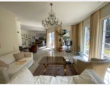 Wohnzimmer mit heller Einrichtung, großen Fenstern und beigen Vorhängen. Im Vordergrund befinden sich ein Glastisch, ein klassischer Teppich und weiße Sofas, während der Raum in den Essbereich mit Esstisch und Stühlen übergeht. Ein Kronleuchter hängt von der Decke, und Pflanzen sowie ein Fernseher sind ebenfalls sichtbar.