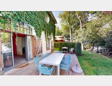 Außenbereich eines Gebäudes mit efeubewachsener Fassade, bodentiefen Fenstern und Holztüren. Eine Holzterrasse mit einem langen Tisch, blauen Stühlen und einem kleinen Grill grenzt an eine Hecke und eine grüne Wiese, die von Bäumen umgeben ist.