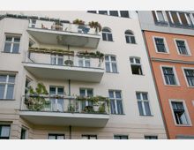 Mehrstöckiges Wohngebäude mit hellen Fassaden, zahlreichen rechteckigen Fenstern und mehreren Balkonen, die mit Pflanzen und Möbeln dekoriert sind. Rechts daneben ein angrenzendes Gebäude mit orangefarbener Fassade.