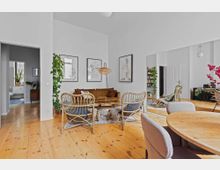 Wohnzimmer mit hellem Holzboden, weißen Wänden und moderner Einrichtung. Sichtbar sind ein Ledersofa, Rattanstühle, ein runder Holzesstisch mit gepolsterten Stühlen, Pflanzen, Wandbilder und ein großer Spiegel, der den Raum reflektiert.