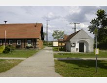 Historische ländliche Szene mit einem Fachwerkhaus links, einem kleinen weiß verputzten Gebäude mit Satteldach rechts und einem Gehweg in der Mitte. Im Hintergrund sind ein Schaukasten, Bäume und Stromleitungsstrukturen zu sehen, zudem ein Haltestellenschild im Vordergrund.