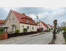 Wohnstraße mit Einfamilienhäusern, die rote Ziegeldächer und Putzfassaden in Beige- und Rosatönen haben. Auf der gepflasterten Straße stehen einige geparkte Autos, während der Bürgersteig von gepflegten Vorgärten und geschnittenen Bäumen gesäumt ist.