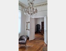 Heller Altbau-Innenraum mit hohen Decken, Stuckfries und großem Kristalllüster über Holzdielenboden. Offene doppelflügelige Tür führt in ein angrenzendes Zimmer mit Vitrine und kleinem Tisch; an der Wand hängt ein goldgerahmter Spiegel.