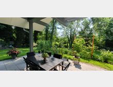 Überdachte Terrasse mit einem dunklen Tisch und mehreren Stühlen, umgeben von Pflanzen in Töpfen. Dahinter ein Garten mit gepflegtem Rasen, Bäumen und dichter Vegetation.