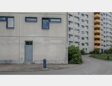 Ein Wohnblock aus Beton mit grauer Fassade, kleinen, rechteckigen Fenstern und gelben Balkonen. Vor dem Gebäude befindet sich ein Gehweg, eine Straße sowie etwas Grünfläche mit Büschen und Rasen.
