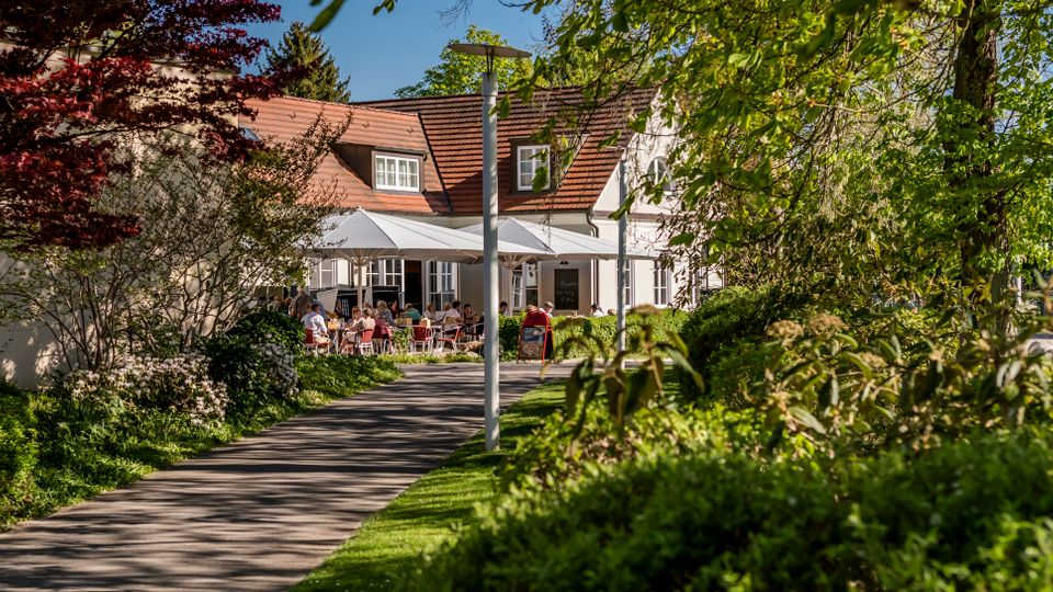 Paved garden walkway leading to a white building with a red tiled roof and dormer windows, set among lush trees and shrubs. In front of the building, large white patio umbrellas cover an outdoor terrace seating area.
