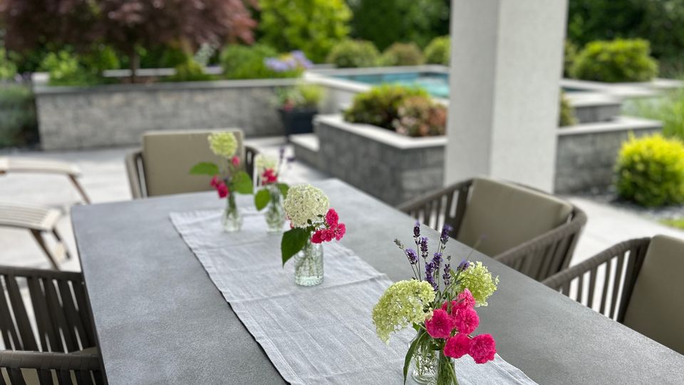 Überdachte Terrasse mit einem langen Esstisch und gepolsterten Gartenstühlen; auf einem weißen Tischläufer stehen mehrere kleine Glasvasen mit Blumen. Im Hintergrund liegt ein modern gestalteter Garten mit niedrigen Steinmauern, Beeten und einem Pool.