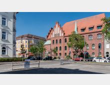 Straßenszene mit historischen Gebäuden: Rechts ein großes rotes Backsteingebäude mit grünen Fenstern und einem spitz zulaufenden Turmdach; links ein cremefarbenes Wohnhaus mit Verzierungen und daneben ein gelbes Mehrfamilienhaus mit Balkon. Im Vordergrund geparkte Autos, Bäume und Straßenlaternen.