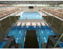 Innenansicht eines Schwimmbads mit einem großen Hauptbecken für Wettkämpfe und einem separaten Sprungbereich mit Sprungbrettern und Plattformen. Auf beiden Seiten befinden sich Tribünen mit Sitzplätzen, über denen ein modernes Dach mit Fenstern für Tageslicht angebracht ist.