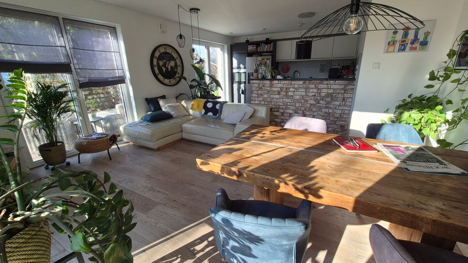 Offenes Wohnzimmer mit moderner, gemütlicher Einrichtung, hellem Holzboden und großen Fenstern mit blickdichtem Rollos. Im Vordergrund ein massiver Holztisch mit Stühlen, im Hintergrund eine Couch, zahlreiche Zimmerpflanzen und eine Küche mit Ziegelwand.