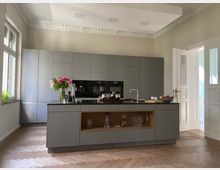 Moderne Küche mit grauen Einbauschränken und einer zentralen Kücheninsel mit integriertem Waschbecken und Stauraum. Ein großer gedeckter Holzfußboden mit Fischgrätmuster ergänzt den Raum, der von hohen Decken mit Stuckverzierungen und großen Fenstern mit natürlichem Licht geprägt ist.