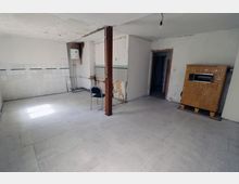 Ein leerer, renovierungsbedürftiger Raum mit blassen Wänden, einer teilgefliesten Wand, freiliegenden Leitungen und einem braunen Kachelofen an der rechten Wand. Ein einzelner Metallstuhl steht in der Mitte des Raums, und ein einfacher Lichtanschluss hängt von der Decke.