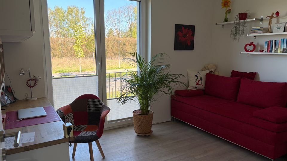 Heller Wohnraum mit großer Glasbalkontür und Blick ins Grüne, einem roten Sofa an der rechten Wand und einer großen Zimmerpflanze davor. Links steht ein kleiner Arbeitsbereich mit Holztisch und Stuhl; an der Wand hängen Regale mit Büchern und Dekoration.