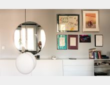 Weiße Kommode mit Schubladen vor einer grauen Wand, an der mehrere gerahmte Bilder in verschiedenen Größen aufgehängt sind. Über der Kommode hängt ein runder Spiegel, der ein Fenster und eine Person mit Kamera reflektiert, sowie eine kugelförmige Hängelampe im Vordergrund.