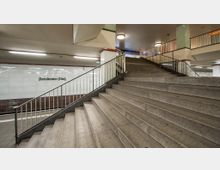 U-Bahnstation mit einer breiten Treppe, die zu einer oberen Ebene führt, umgeben von schlichten Metallgeländern. Im Hintergrund ist eine mit weißen Fliesen verkleidete Wand zu sehen, auf der „Potsdamer Platz“ steht.