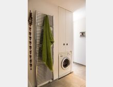 Kleine Waschküche mit glänzendem Heizkörper, an dem ein grünes Handtuch hängt. Rechts befindet sich eine eingebaute Waschmaschine unter weißen Schränken, daneben eine helle Wand mit einem kleinen Foto.