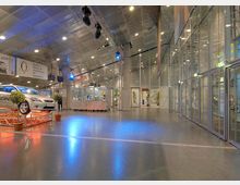 Großer, moderner Innenraum mit hohen Decken und Glaswänden; auf der linken Seite steht ein Auto auf einer Ausstellungsplattform, umgeben von kleinen Pflanzen in Töpfen. Im Hintergrund befinden sich Infotafeln, eine beleuchtete Theke und eine offene, helle Atmosphäre mit deckenmontierten Scheinwerfern.