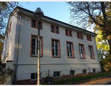 Zweistöckiges Gebäude mit cremefarbener Fassade und rot umrahmten Fenstern, umgeben von Bäumen und einem Zaun. Im Vordergrund steht eine altmodische Straßenlaterne, und der Himmel ist klar und blau.