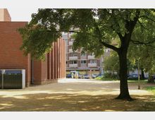 Ein offener Platz mit einem großen Baum im Vordergrund und einem Gebäude aus rotem Backstein auf der linken Seite. Im Hintergrund sind Wohnhäuser, geparkte Autos und zwei Personen neben einem Fahrrad zu sehen.