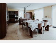 Moderne, helle Küche mit weißen Wänden und großen Fenstern, ausgestattet mit Holzeinbauküchen, Edelstahlgeräten und einer zentralen Kücheninsel. Im Vordergrund steht ein großer Esstisch mit weißen Tischdecken, dekoriert mit einem Blumenarrangement.