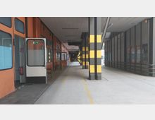 Industriehalle mit einem überdachten Gang, links orangefarbene Wände mit Fenstern und einer offenen Tür, rechts Stahlgeländer und graue Säulen mit gelb-schwarzen Markierungen. Der Boden ist aus Beton und durch gelbe Linien markiert.