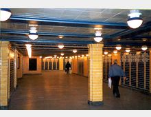 Unterirdischer Korridor mit gelben Fliesenwänden, blau lackierten Stahlträgern und mehreren kugelförmigen Deckenlampen. Personen gehen durch den beleuchteten Durchgang, der zu einer Eingangstür oder einem weiteren Bereich führt.