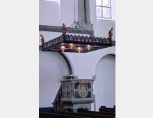 Innenraum einer Kirche mit einer verzierten hölzernen Kanzel und einem hängenden Kronleuchter in Form eines rechteckigen Rahmens mit Figuren an den Ecken. Die Wände sind weiß, mit Rundbögen und hohen Fenstern.