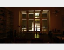 Ein abgedunkelter Speiseraum mit einem großen Holztisch in der Mitte, dekoriert mit Vasen und Kerzenhaltern. Durch die großen Fenster ist bei Nacht ein beleuchtetes Gebäude mit klassischer Architektur sichtbar.