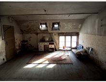 Ein leerer, alter Dachbodenraum mit abgenutzten Wänden und Holzboden. Im Raum stehen ein kleiner Tisch mit einem Stuhl, ein Sofa, ein Teppich, ein altes Waschbecken und eine kleine Kommode; Licht fällt durch das Fenster und zwei kleine Dachfenster.
