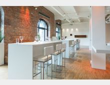 Innenraum eines modernen Veranstaltungsortes mit Backsteinwänden und großen, halbkreisförmigen Fenstern. Zu sehen sind hohe weiße Tische mit passenden Barhockern sowie gedeckte Tische mit Gläsern und Dekoration auf einem Holzboden.