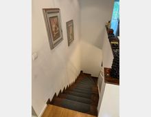 Ansicht eines modernen Treppenhauses in einem Wohngebäude mit Holztreppenstufen, die mit dunklen Teppichauflagen versehen sind. An der weißen Wand links hängen zwei gerahmte Bilder; rechts befindet sich ein Treppengeländer sowie eine Ablage mit persönlichen Gegenständen.