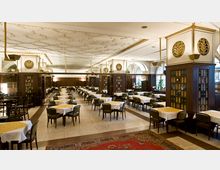 Großer, eleganter Speisesaal mit symmetrisch angeordneten Tischgruppen, weiß eingedeckt mit gelben Tischläufern. Der Raum ist durch dunkle Holzelemente, dekorative Verzierungen und ein detailreiches, helles Deckendesign geprägt.