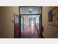 Langer Innenflur eines Schul- oder Bürogebäudes mit roten Linoleumboden, beigen Wänden und Deckenleuchten, durch eine Glastür mit darüberhängendem Notausgangsschild. Rechts verläuft eine Fensterreihe mit Heizkörpern, daneben hängen Aushänge; an der Wand ist ein Feuerlöscher montiert.