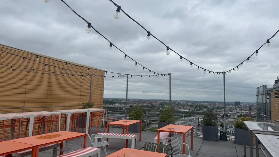Rooftop terrace dining area with orange tables, striped benches, and metal chairs beneath string lights, bordered by railings and planter boxes. The terrace overlooks a wide city skyline under an overcast sky, with a wood-paneled wall on one side.