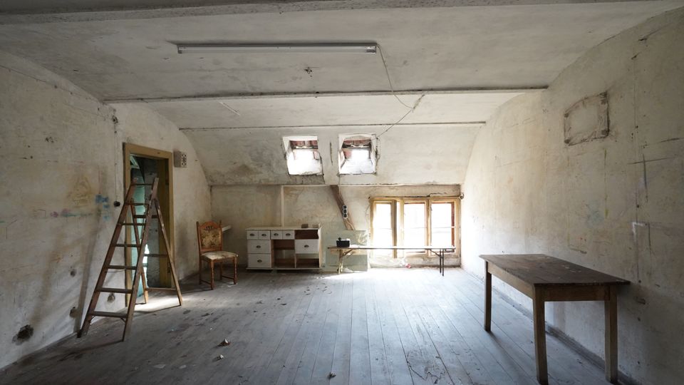 Großer, leerer Dachbodenraum mit schrägen Decken, abgenutztem Holzboden und fleckigen, teils rissigen Wänden. Hinten sorgen kleine Dachfenster und ein größeres Fenster für Licht; im Raum stehen eine Leiter, ein Tisch sowie einige alte Möbel.