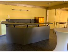 Gelber Raum mit einer modernen, dunklen Theke und einer Edelstahlspüle, einem gelben Retro-Kühlschrank im Hintergrund und einer runden Stehtischhusse im Vordergrund. Im Raum sind gelbe Wände, ein dunkler Boden und ein Geländer sichtbar.