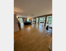 Großzügiges Wohnzimmer mit hellem Holzfußboden und großen Fenstern, die viel Tageslicht hereinscheinen lassen. Der Raum ist mit einem grünen Sofa, einem Esstisch mit Stühlen und diversen dekorativen Elementen ausgestattet, und grenzt an eine Terrasse mit Blick ins Grüne.