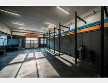 Ein moderner Trainingsraum mit hohen Decken und schwarzen Gummibodenmatten, ausgestattet mit Metallgerüsten für Klimmzüge, Turnringen und Scheibenstapeln an der Wand. Die Wände sind dunkelgrau mit einer orange-blauen Linie, und große Fenster lassen Tageslicht herein.