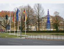 Kreuzung mit Blick auf eine blaue Glas-Skulptur in Form eines Turms, flankiert von mehreren nationalen und europäischen Flaggen an Fahnenmasten. Im Hintergrund stehen kahle Bäume, ein grüner Parkbereich und Gebäude mit Schrägdächern.