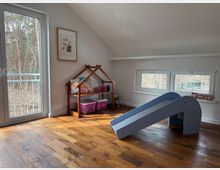 Bright attic playroom with a sloped ceiling, hardwood floor, and large windows and a glass door overlooking trees. The room includes a small indoor slide and a wooden house-shaped shelf with storage bins against a light-colored wall.