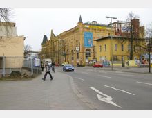 Straßenszene mit einem großen Backsteingebäude mit dem Schriftzug „Südhaus“ und mehreren Werbeplakaten. Im Vordergrund ist eine mehrspurige Straße mit Fußgängern, Autos und Radfahrern zu sehen, flankiert von Gehwegen und einigen Bäumen.