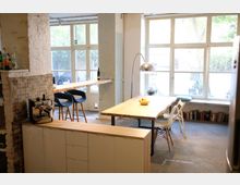 Heller Küchen- und Essbereich mit großen Fenstern, Holzmöbeln und Betonboden. Im Vordergrund eine Küchenzeile mit Kaffeemaschine, im Hintergrund ein Esstisch mit Stühlen und ein Bartresen mit Hockern.