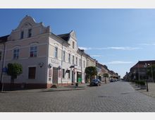 Straßenszene in einer Kleinstadt mit gepflasterter Fahrbahn und schmalen Gehwegen, umgeben von zweigeschossigen historischen Gebäuden mit heller Fassadengestaltung. Links befindet sich ein Eckhaus mit der Aufschrift "Ratskeller", während entlang der Straße kleine Bäume und geparkte Autos sichtbar sind.