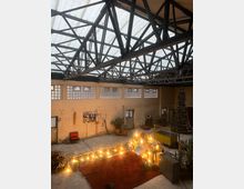 Eine alte Industriehalle mit sichtbaren Stahlträgern unter einem lichtdurchlässigen Dach. Der Raum ist teilweise dekoriert, mit einer Lichterkette um einen Sitzbereich auf einem roten Teppich, Pflanzen, einem geschmückten Weihnachtsbaum und verschiedenen Möbelstücken.