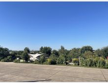 Weitläufige Betonfläche im Vordergrund mit angrenzendem Gartenbereich voller Bäume, Sträucher und Blumen; im Hintergrund sind kleinere Gebäude mit weißen, zeltartigen Dächern zu sehen, umgeben von grünem Bewuchs, unter einem klaren blauen Himmel.